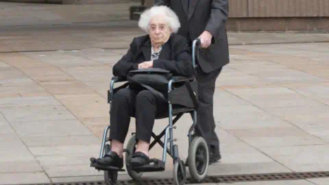 PA Media June Mills, with white hair and wearing a black blazer and black trousers, sits in a wheelchair and clutches her handbag as she is pushed away from Liverpool Crown Court by her husband 