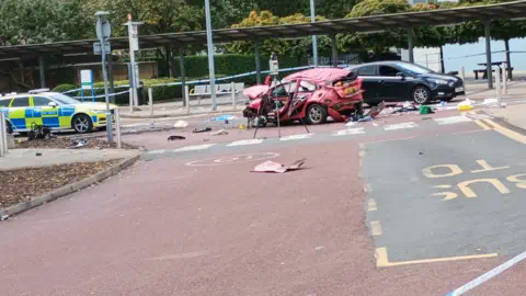 @GrantTheHatter Mangled wreckage of red car surrounded with debris (middle) and police car (left) with a black car behind the crashed one