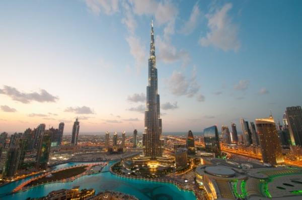 A Dubai cityscape is pictured at sunset. The city is illuminated in the bottom portion of the frame by synthetic light, as natural light lights the top half of the photo. A modern skyscraper points up into the sky in the center of the frame. rare view from the address hotel which is no longer possible.