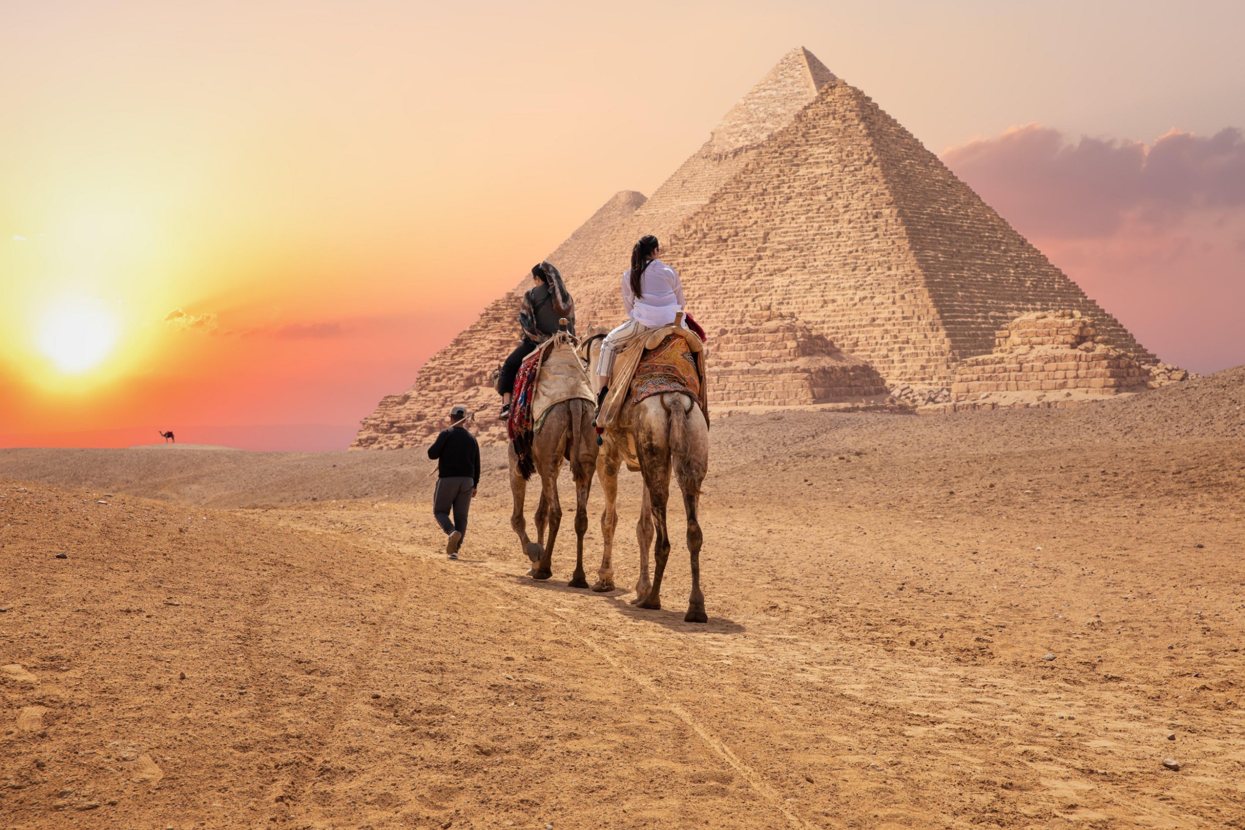 Tourists on camels near the Great Pyramids of Giza, Egypt, with the setting sun casting red light on the horizon.