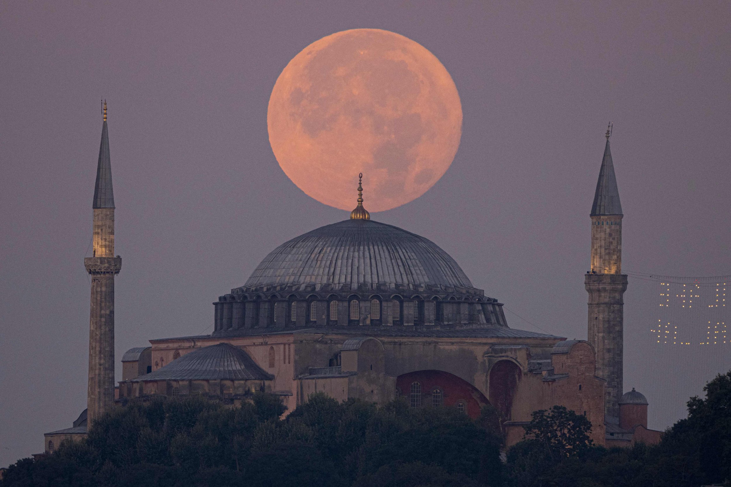 The red Sturgeon super moon rises in a hazy purple sky over the domed roof and pointed minarets of Ayasofya-i Kebir Camii or Hagia Sophia Grand Mosque in Istanbul.
