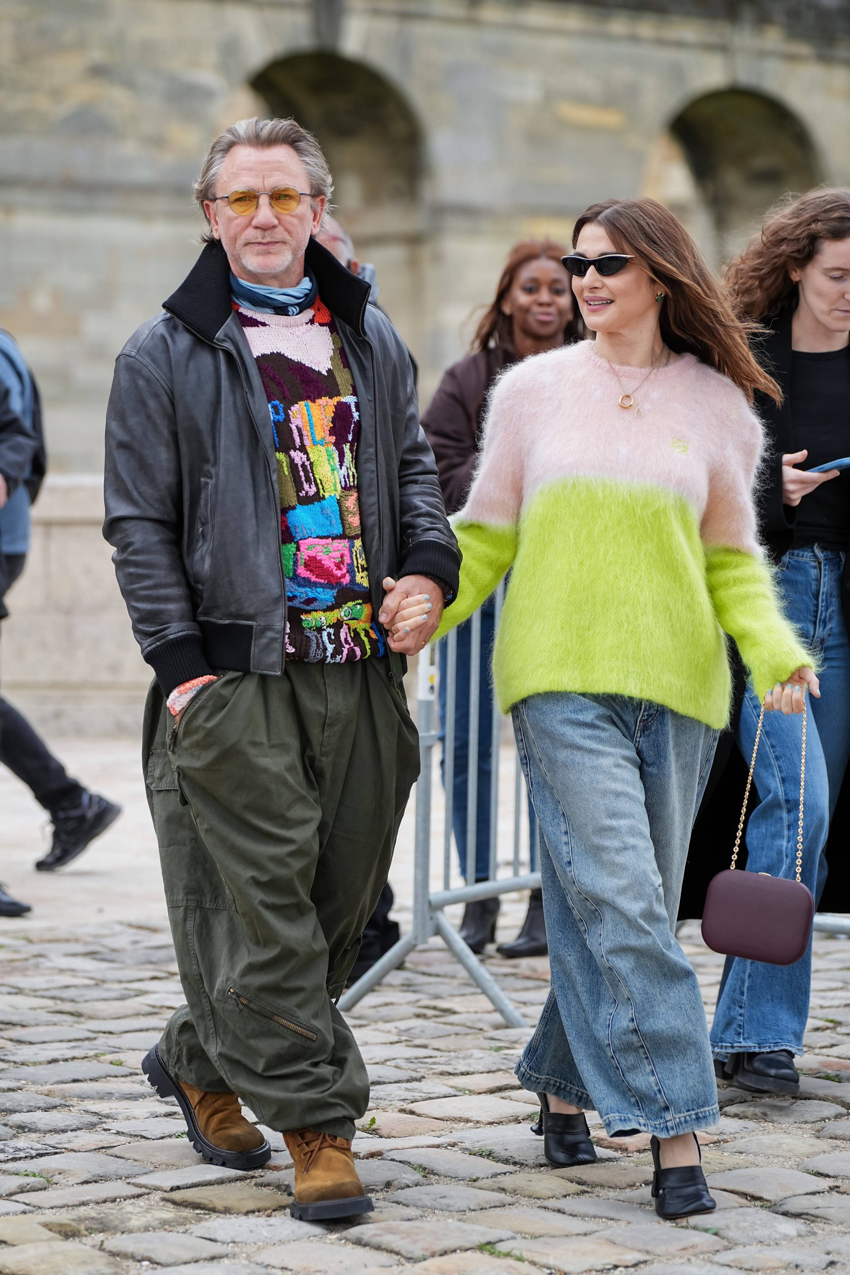 Daniel Craig and Rachel Weisz at the Loewe fashion show. He is wearing a black bomber jacket, unusual patterned jumper and khaki trousers. She is wearing a pink and yellow jumper and a baggy blue jeans