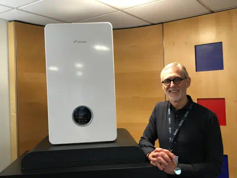 A man wearing black-rimmed glasses, a black shirt and company lanyard sits next to a plinth in an office, with a boiler sitting on it.