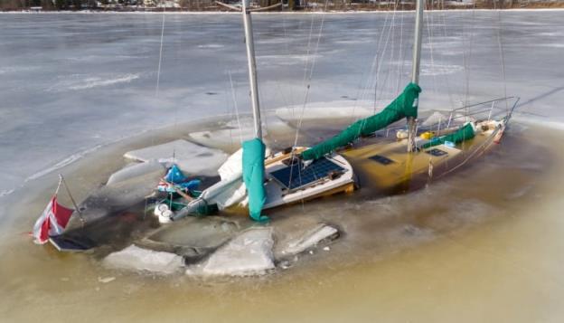 Double-masted sailboat submerged in ice nearly to the top of the roof of the cabin and surrounded by a circle of brownish water and then white ice outside the circle.