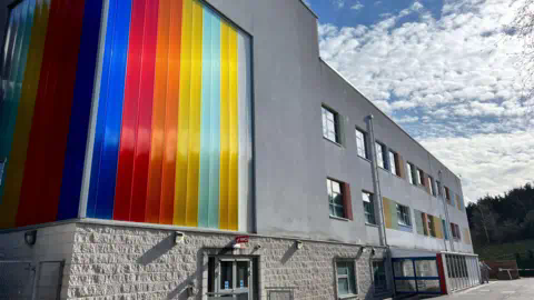A grey hospital building with a window covered in a multi-coloured rainbow panelling.