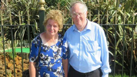 家庭资料:Lois and John McCullough,他们站在一个迷你高尔夫球场前。他们穿着蓝色衣服,对着摄像头微笑。