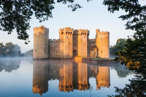 Bodiam Castle surrounded by its misty moat seen through surrounding trees. A near-perfect reflection is made in the moat water.