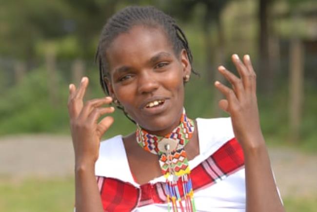 A young African woman with a beaded necklace smiles and gestures at the camera