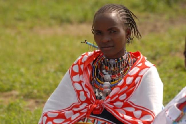 A young African woman with a beaded necklace, braided hair and a cloth over her shoulders looks at the camera