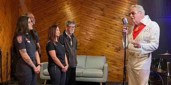 Four people listening to an Elvis Presley impersonator recording music in a studio, wearing a classic white suit and holding a microphone.