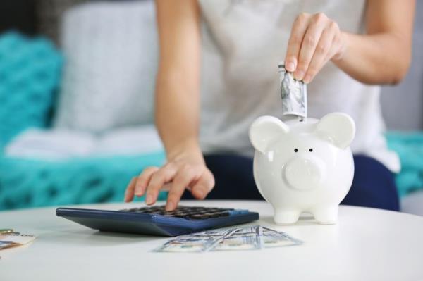 A woman using a calculator drops cash into a piggy bank
