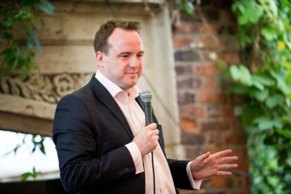 Matt Forde speaking into a microphone while wearing a black suit jacket and white shirt