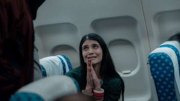 A woman begging to a masked man while sitting in a plane seat in a scene from IC 814: The Kandahar Hijack.