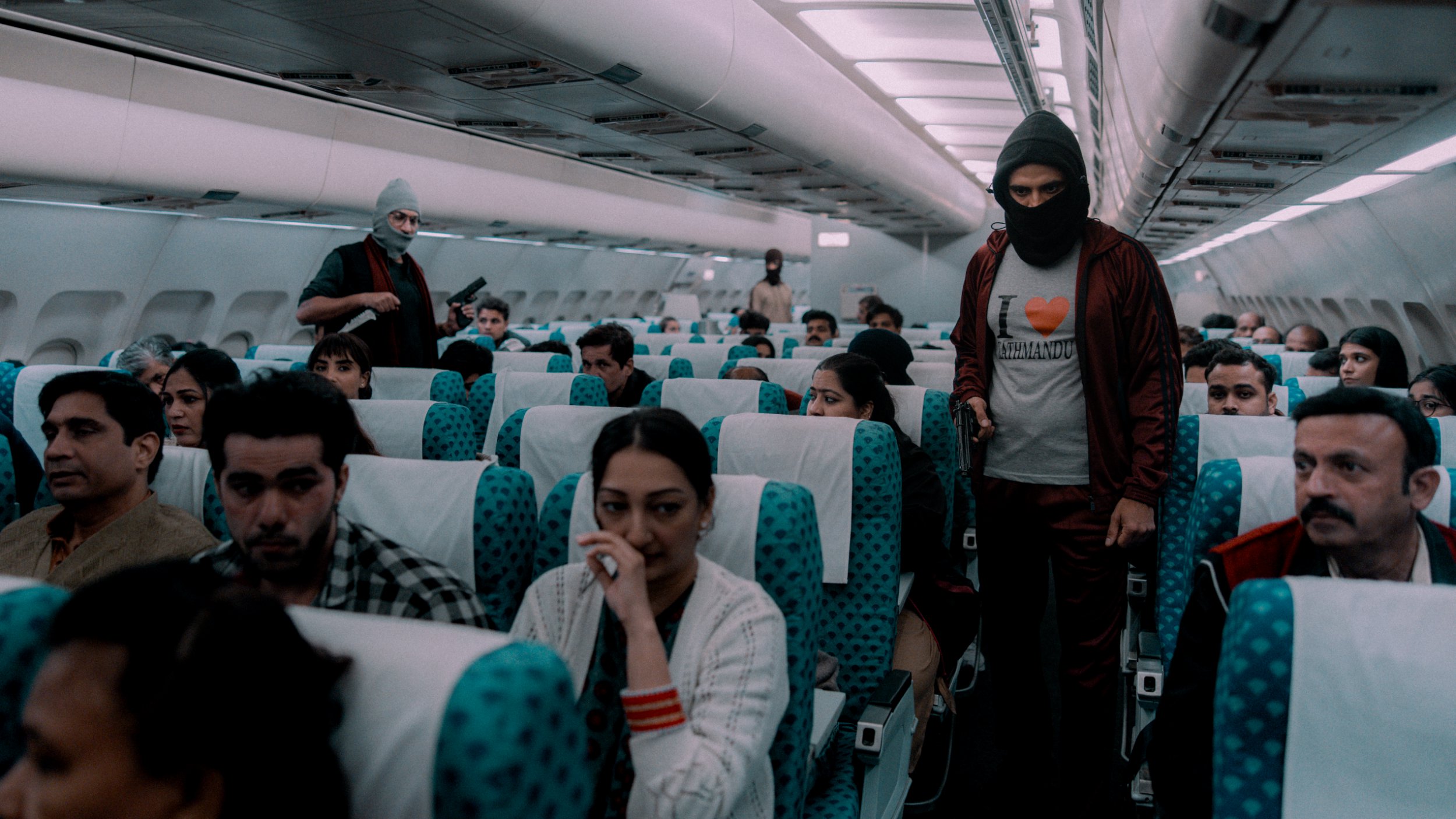 A scene from IC 814: The Kandahar Hijack showing a masked man walking down the aisle of an airplane holding a gun.