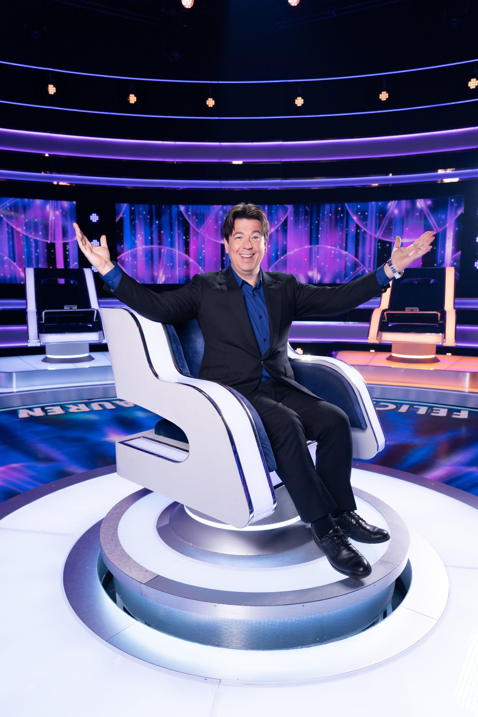 Michael McIntyre sits in The Wheel chair with his arms raised up