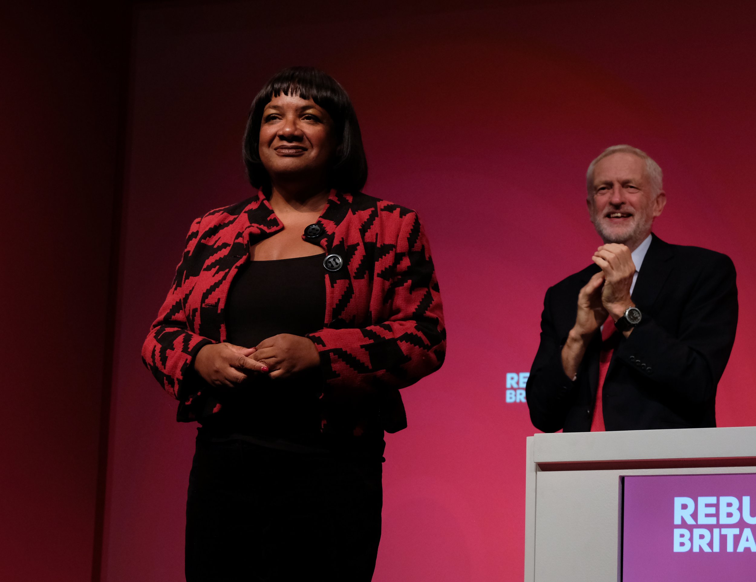 Diane Abbott stands on stage with Jeremy Corbyn applauding her from behind