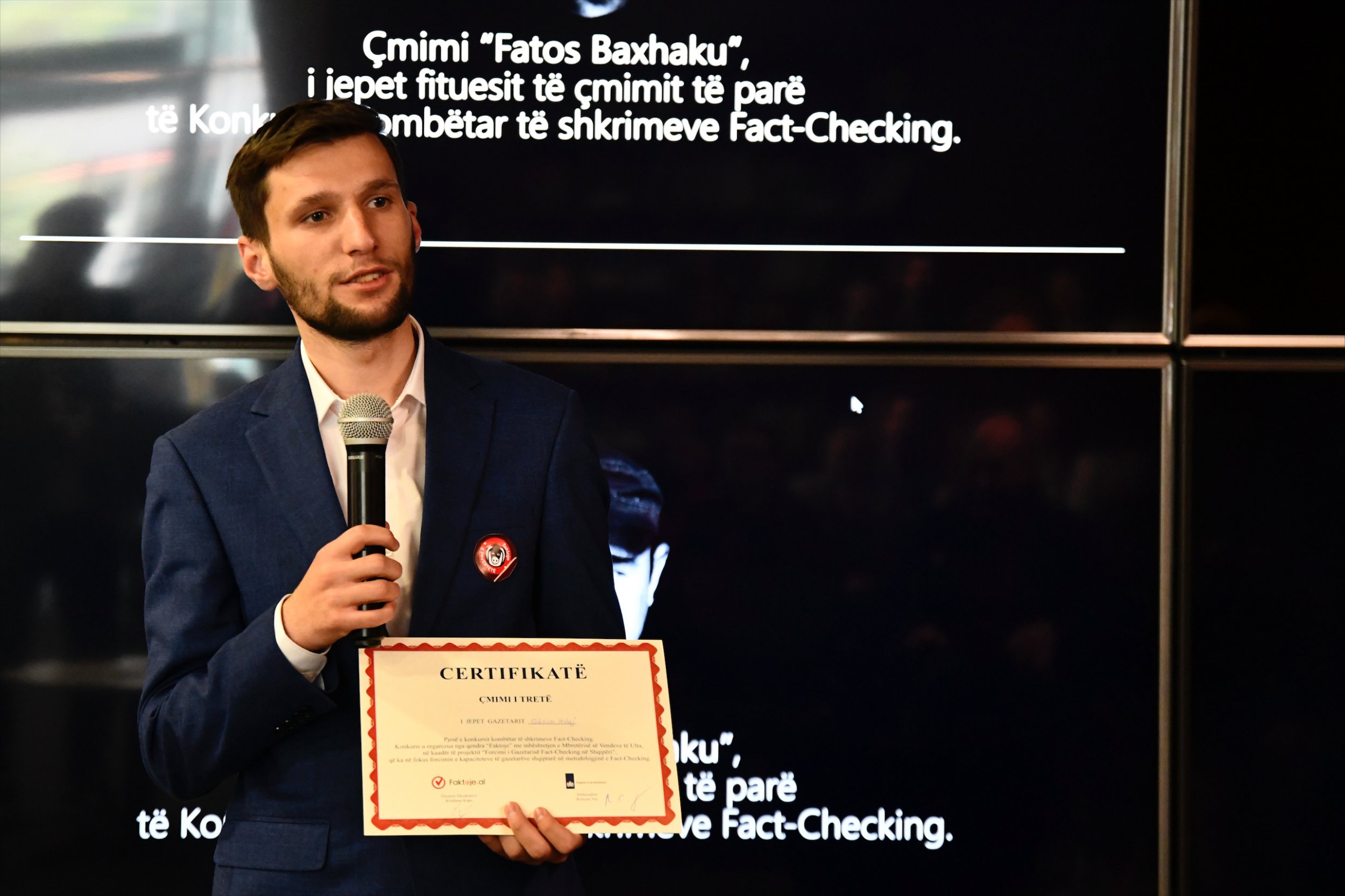 Gezim holding a certificate and microphone, in front of a screen that has words in Albanian on ot