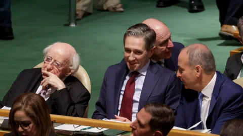 BBC Three men, Michael D Higgins on the left wearing a black suit, white shirt and pink tie. In the middle is Taoiseach Simon Harris in a navy suit, blue shirt and blood red tie. In a navy suit, white shirt and blue tie on the right is Tánaiste Micheál Martin