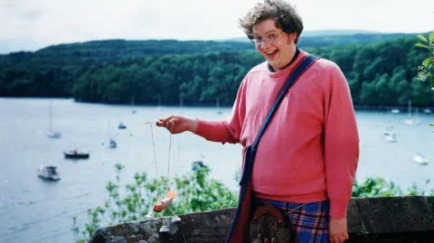 Miles Jupp, as Archie the inventor in Balamory, stands next to a low wall overlooking the harbour at Tobermory, holding a puppet made with egg boxes and wearing a pink jumper and kilt with sporran.