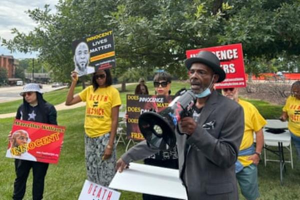 man wearing black hat and gray suit speaks into speakerphone as people around him hold signs