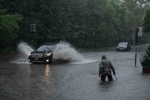 Man wades through thigh-high waters during last week's flooding to clear storm drains as car passes, shooting up water alongside it as it goes through flooded waters.