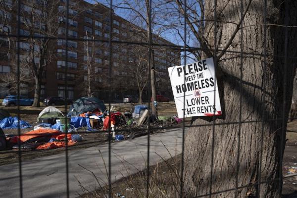 A sign that says 'Please help the homeless: lower the rents' is seen on a tree beside a group of tends sent up in a city park