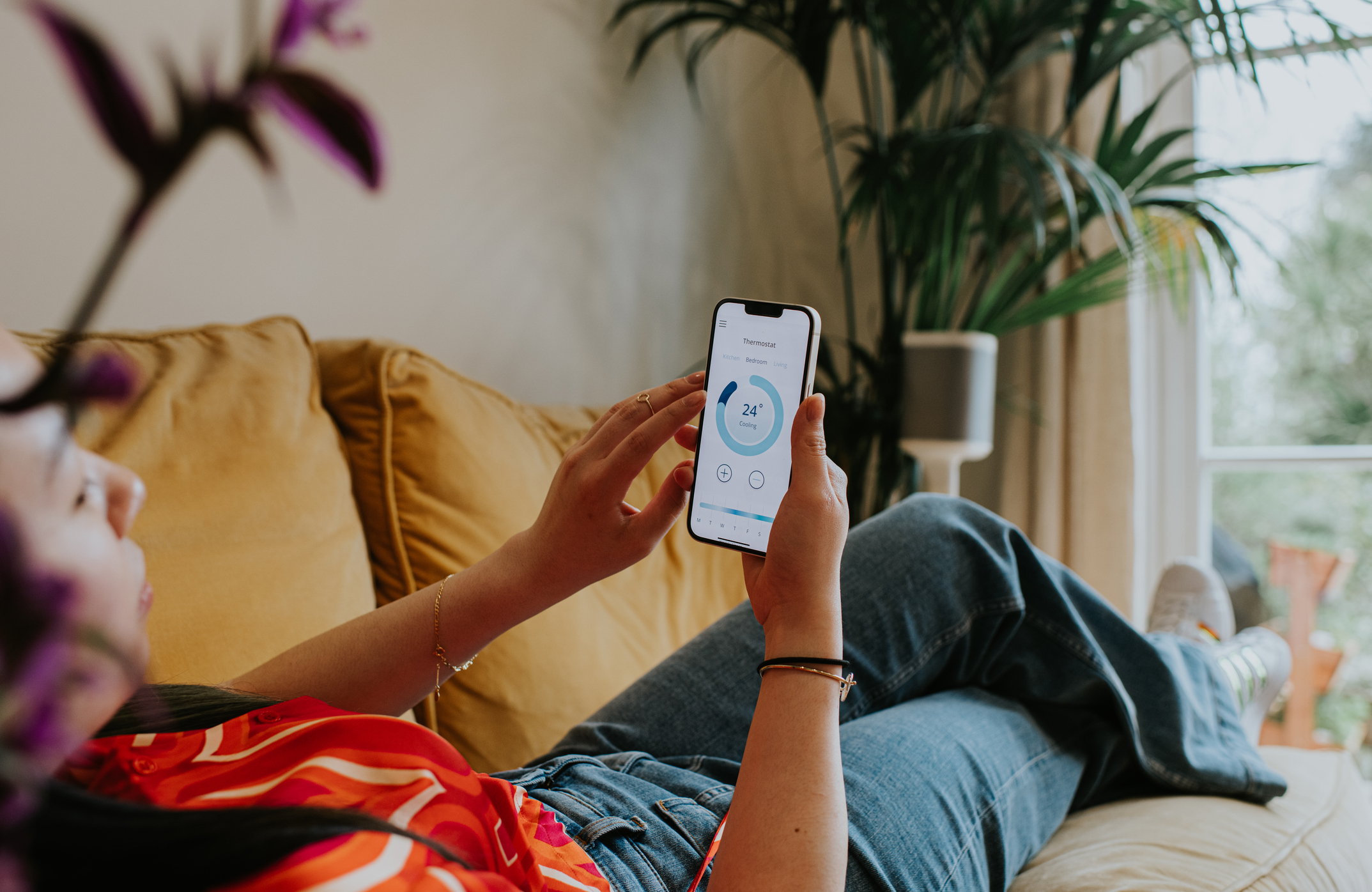 Relaxed woman reclines on a sofa and uses a thermostat app on her smart phone to control the housing heating system