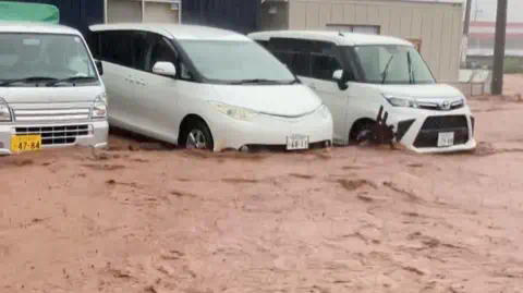 NHK 中部日本一条道路上涌现出褐色洪水。三辆白色车辆停放在水中,水淹到车轮和保险杠