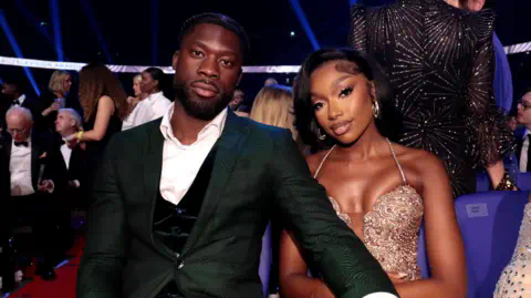 Getty Images Josh Oyinsan and Mimi Ngulube during the NTAs. Josh wears a black tuxedo with waistcoat and unbuttoned shirt, his hand on Mimi's knee. Mimi wears an embellished nude dress. They're pictured at the event with a crowd seated behind them.