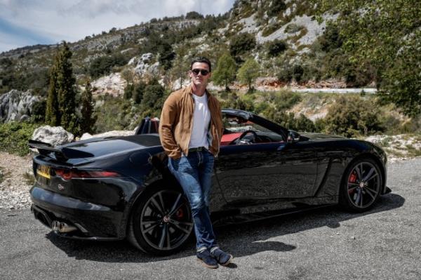 Luke Evans in jeans, a white T-shirt, brown jacket and shades leans against a black Jaguar F-Type SVR in dramatic mountain scenery