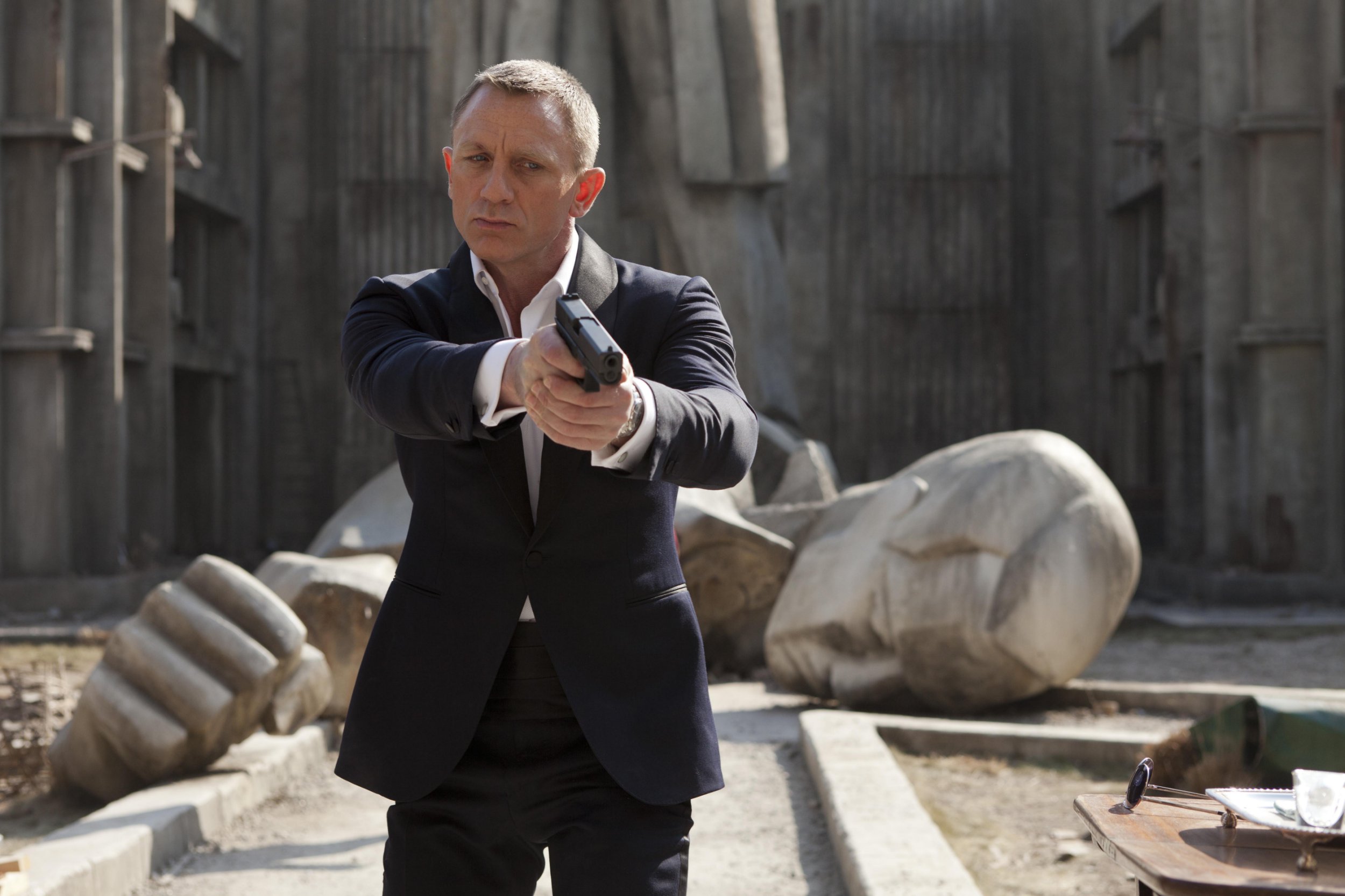Daniel Craig as James Bond points a gun with stone monuments behind him in an outdoors scene from the film Skyfall