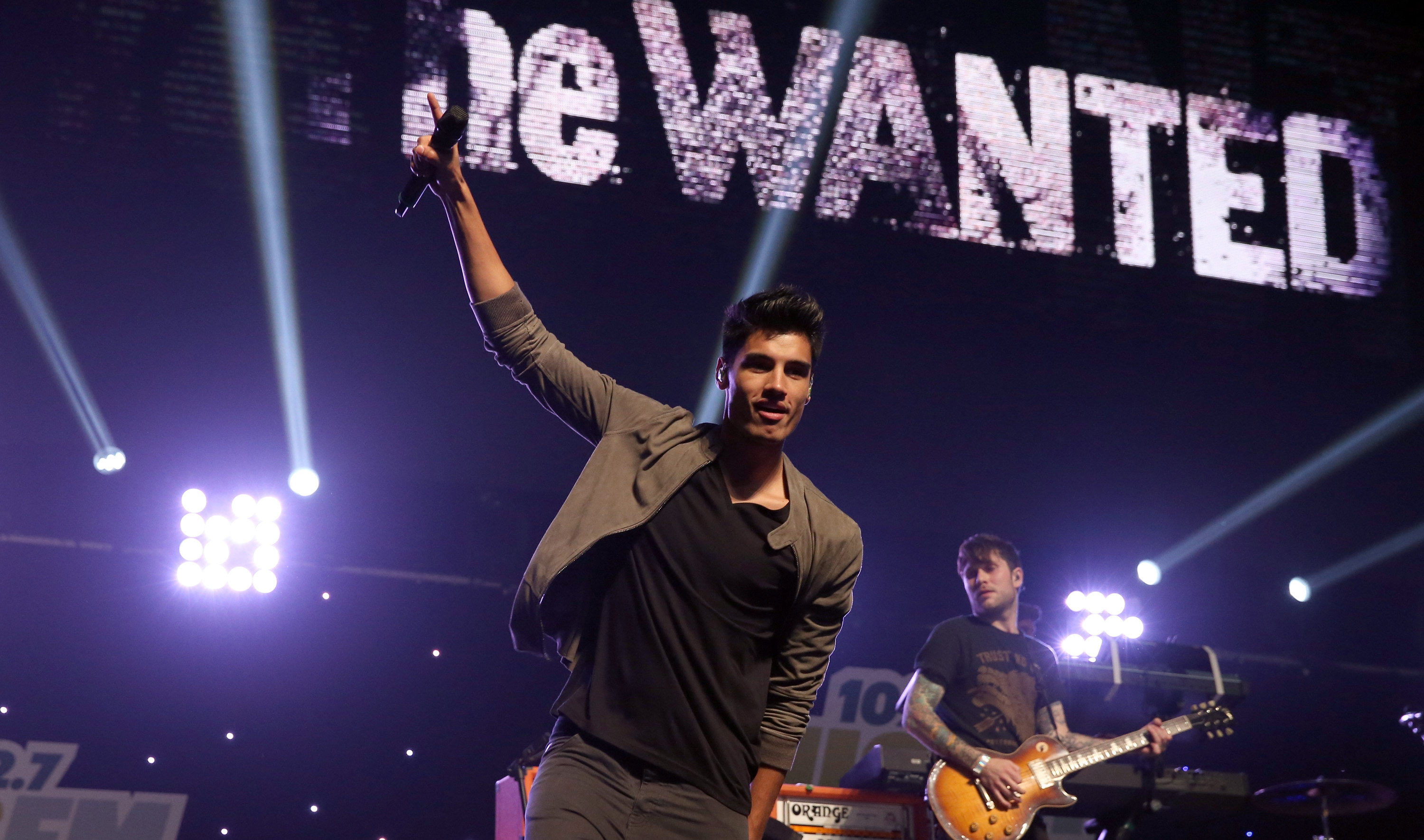 Siva Kaneswaran of The Wanted performing onstage. He is holding a microphone in the air while wearing a black t-shirt and khaki jacket with matching trousers