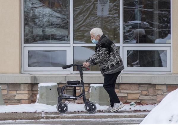 An elderly woman in a knit sweater walks down a sidewalk with the assistance of a walker