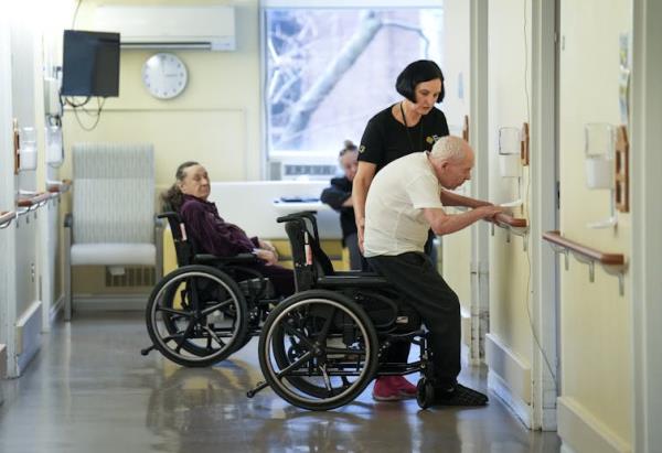 A woman places a hand against the back of an elderly man as he uses a railing to stand up from a wheel chair