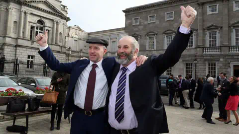 Getty Images Michael Healy Rae (left) with his brother Danny Healy-Rae (right). Both men are wearing suits and have an arm held in the air. Michael is wearing a hat.