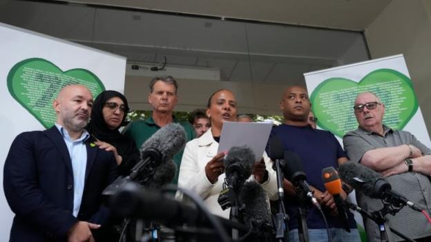A dark-complected woman holds a piece of paper while speaking in front of a bank of microphones. Several other people, men and women, surround her.