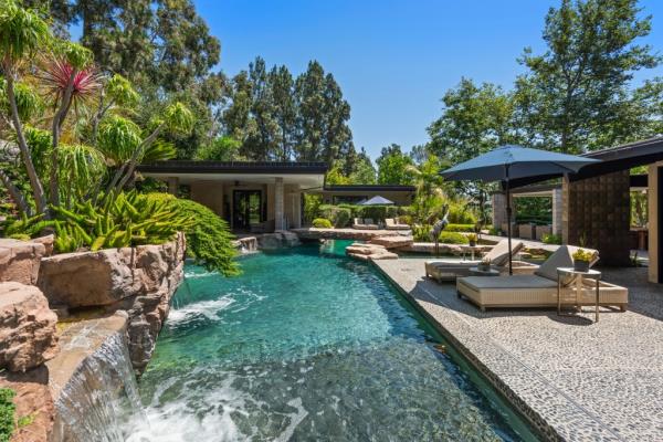 Jeremy Renner's luxurious zen house in Los Angeles with a swimming pool, waterfall, and surrounding trees under a blue sky