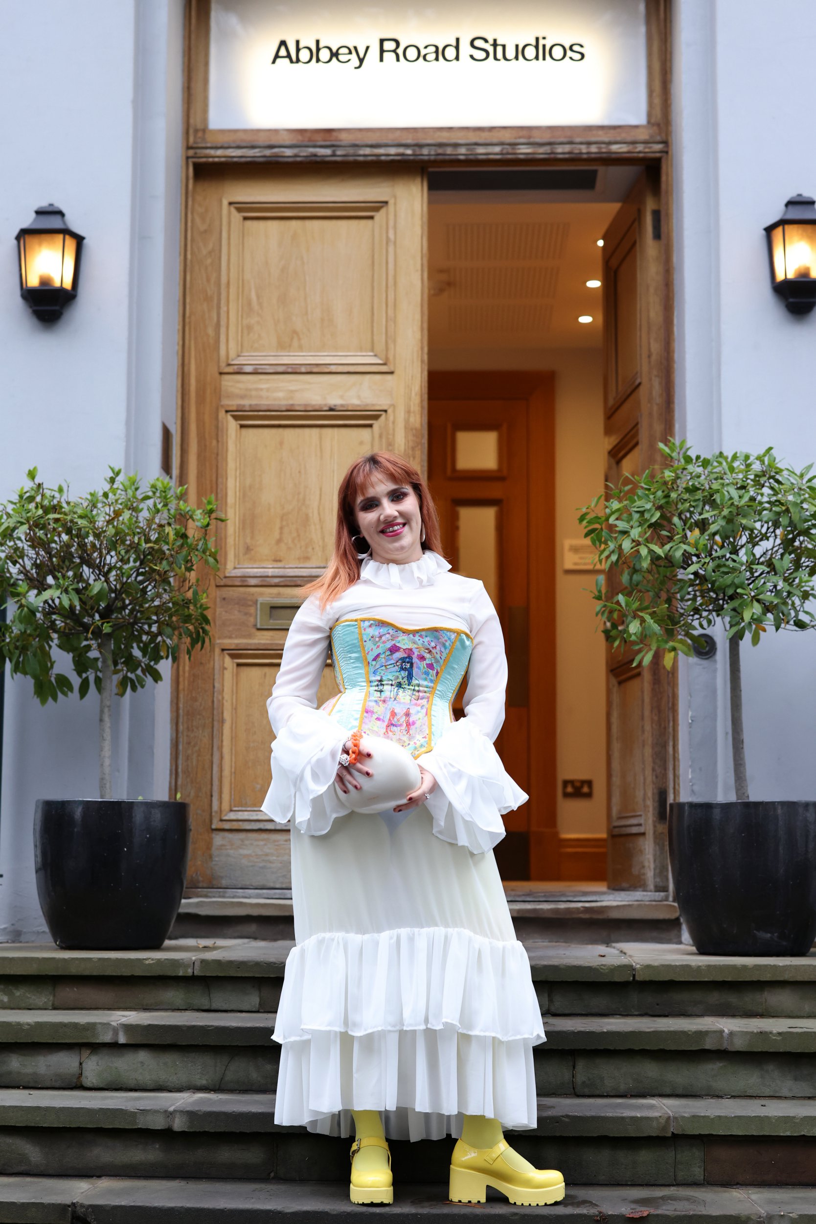 CMAT stands on steps of Abbey Road Studios ahead of the Mercury Music Awards 2024. She wears white frilly dress and corset with her own album artwork on it.