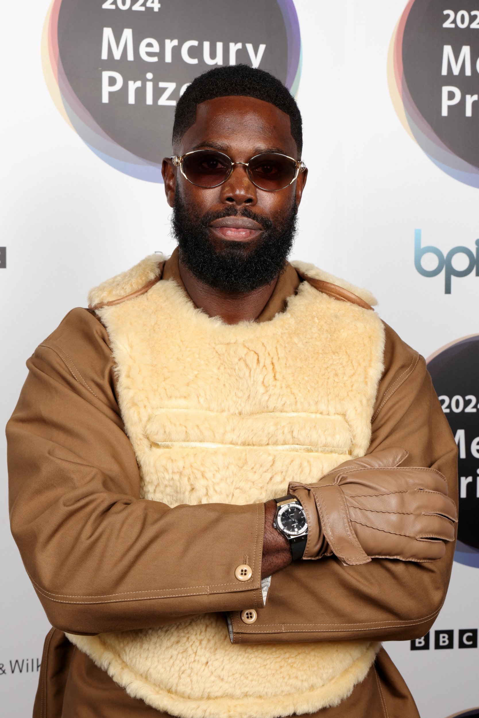 Ghetts arrives for the Mercury Music Awards 2024 at Abbey Road Studios. He wears camel coloured jacket and gloves with fluffy bullet vest over top.