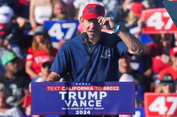 Dennis Quaid speaking at a Donald Trump rally in Coachella, California, wearing a red hat and holding a microphone, on October 12, 2024