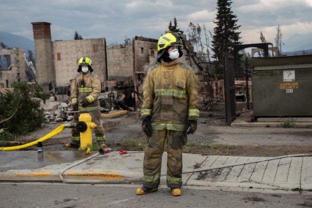 Firefighters in full gear stand before the shell of a burned out building.