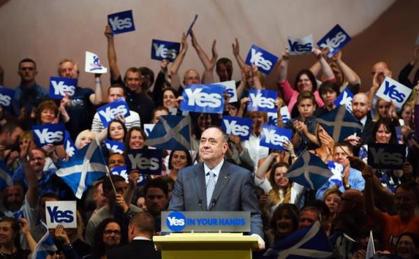 Alex Salmond giving a speech with a crowd of people waving 'Yes' placards behind him.