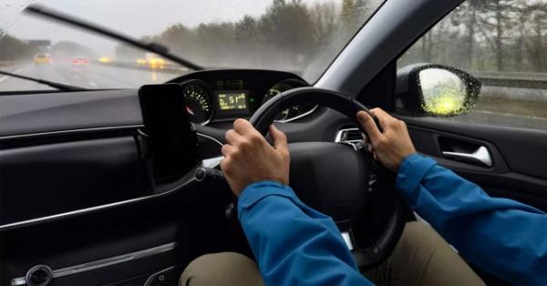 Drivers hands holding a steering wheel with traffic on the road in the distance. 