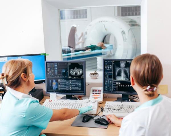 Two nurses look at two computer screens as a patient enters a CT scan machine.