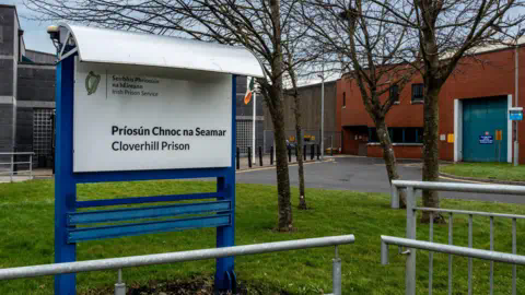 Getty / Noel Bennett A blue and white sign with "Cloverhill Prison" written on it surrounded by grass, metal barriers and trees. In the background is a brown and dark grey building.