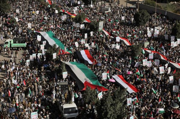 A crowd of thousands marching in the streets, several carrying large Yemeni and Palestinian flags and carrying white signs with Arabic texts.