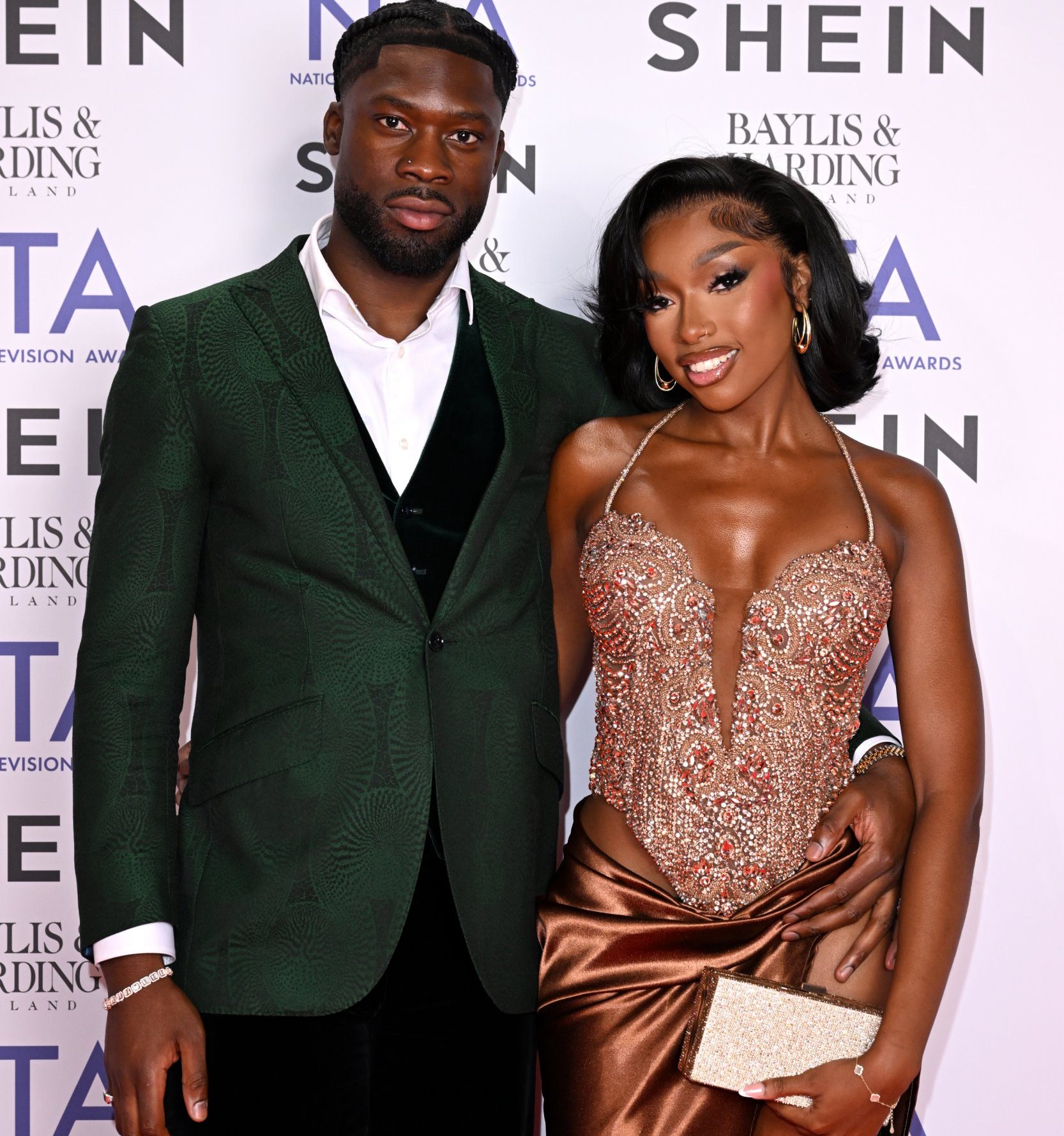 Josh Oyinsan and Mimii Ngulube attend the NTAs 2024. He is wearing a dark green blazer with white shirt, Mimii is wearing a glitzy brown dress. They have their arms around each other's waists and they are smiling.