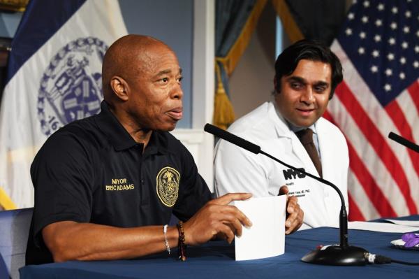 NYC Mayor Eric Adams receiving a Covid shot from Dr. Ashwin Vasan at City Hall, while sitting at a table with a microphone.