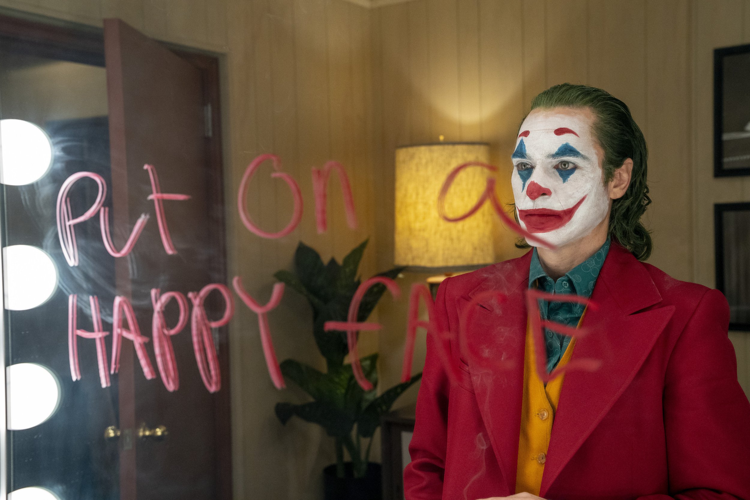 Joaquin Phoenix in a scene from Joker. He looks into mirror wearing clown makeup and suit. 'Put on a happy face' is written on mirror in red lipstick.