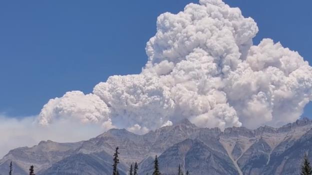 A large smoke plume rises over the Rocky Mountains, under clear blue sky.